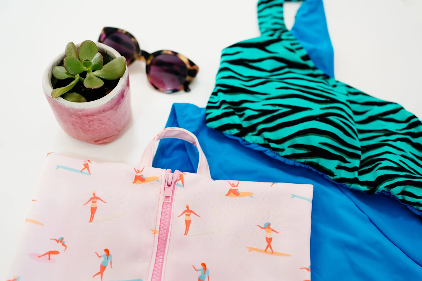 A fabric rectangle featuring a design with small female surfers surfing through a pink background lays on a white surface. A pink zipper has been stitched up the middle. A U-shaped handle in the same fabric has been sewn to the top center of the bag. The