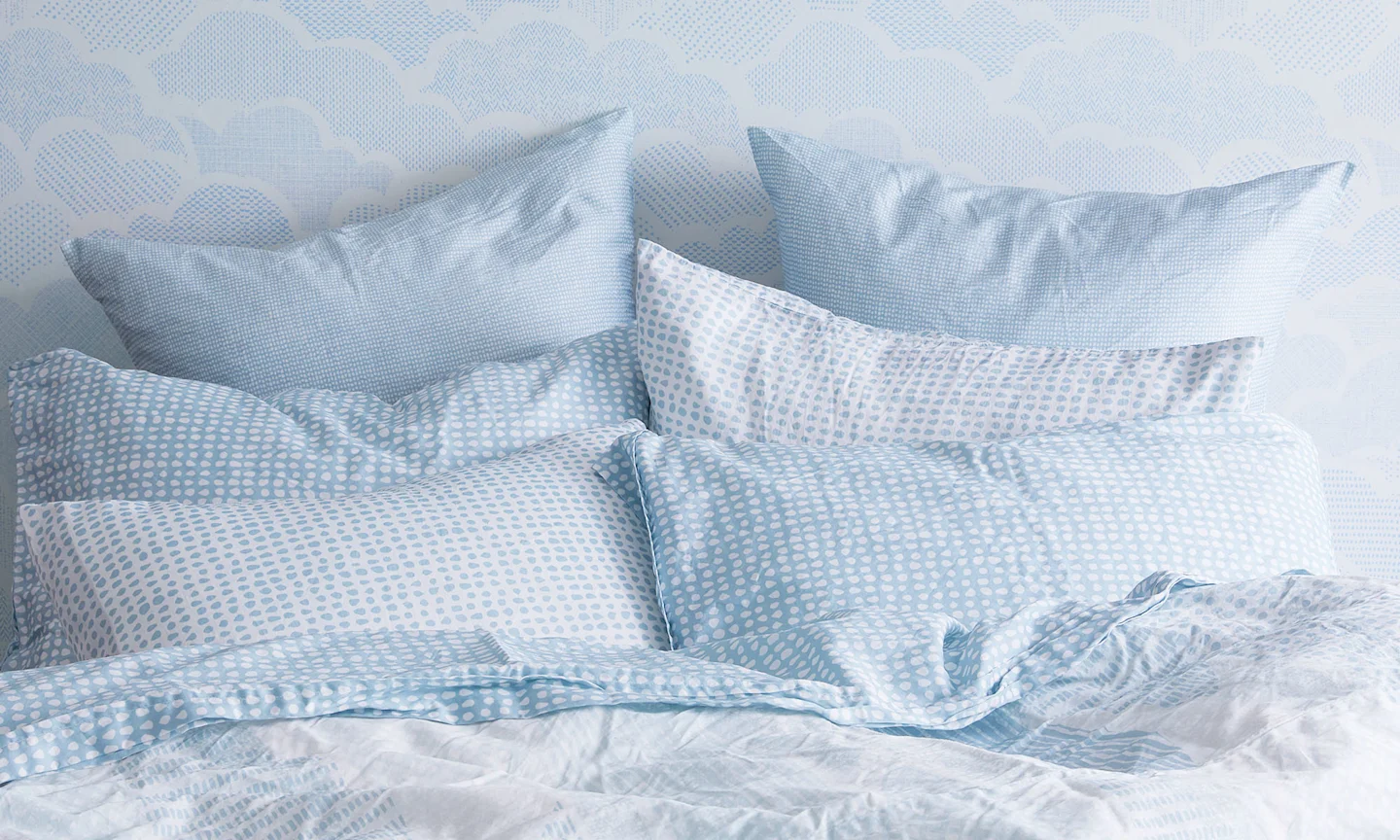 A bed with a soft blue set of pillows shams, sheets, and duvet cover with a coordinating blue cloud wallpaper accent wall.
