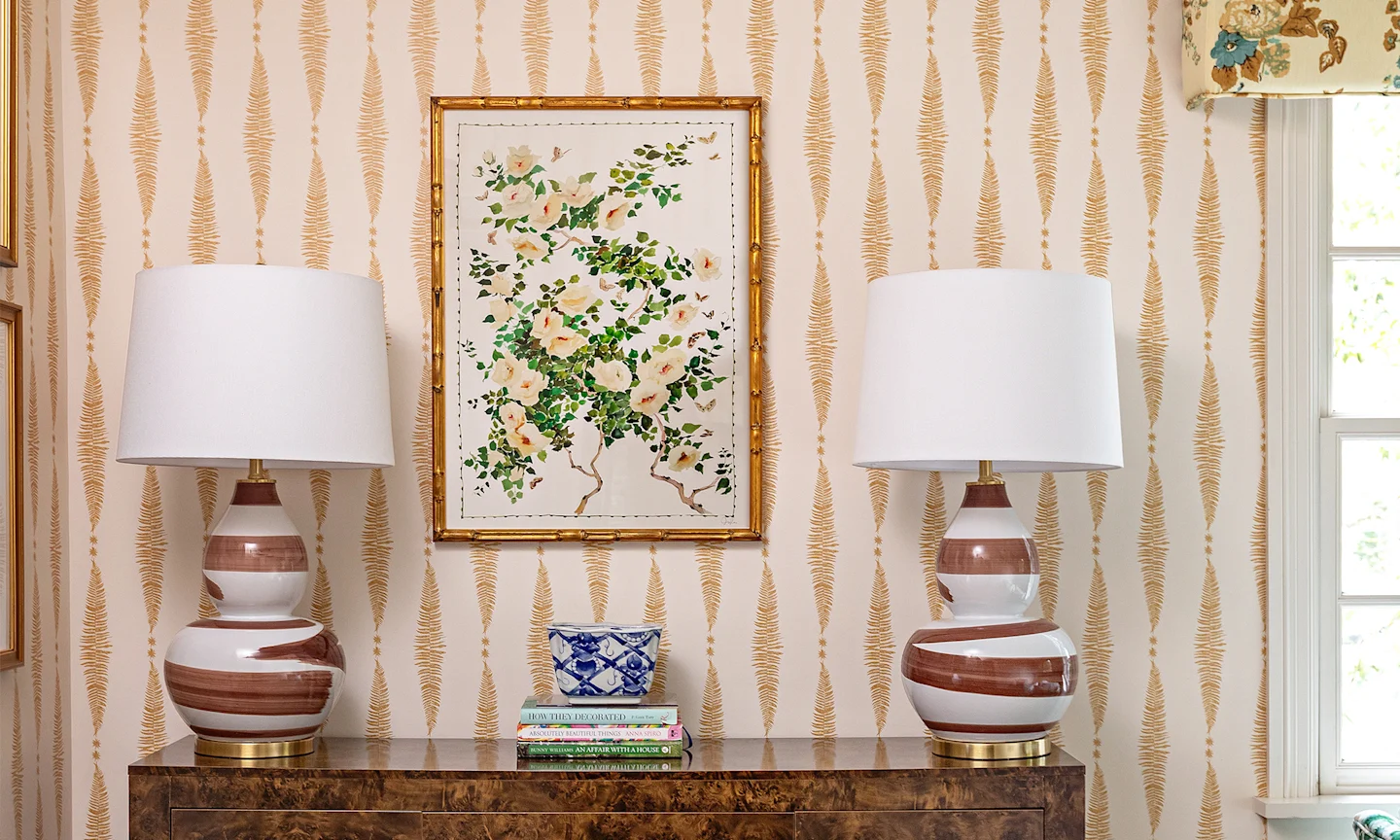 Home office with tam and cream striped wallpaper two lamps and floral art.